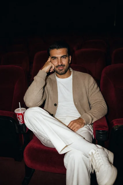 a candid cinematic fashion portrait of a young man sitting alone in an empty movie theater, surrounded by rows of deep red velvet cinema seats, dim moody lighting, flash photography look, relaxed pose with one hand resti…